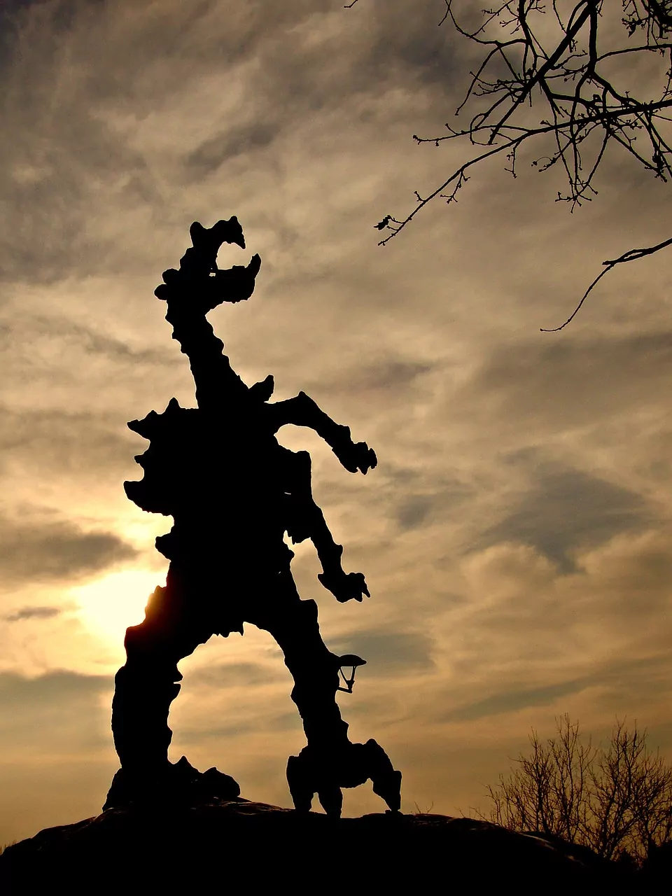 Silhouette of the Wawel Dragon statue with a lamp on its leg, against a cloudy sunset sky. Bare tree branches overhead.