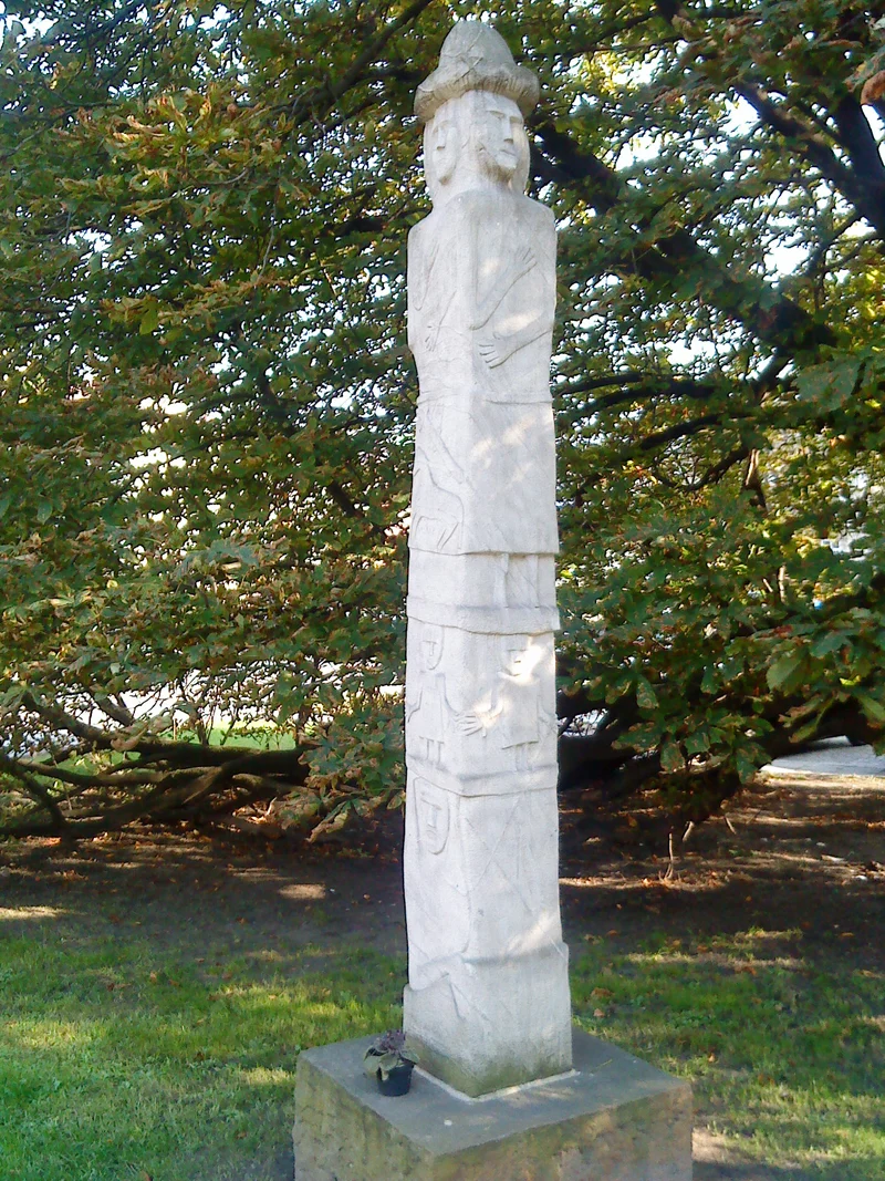 A modern replica of the Zbruch Idol, a tall four-faced Slavic stone pillar, standing in a park in front of a leafy tree.