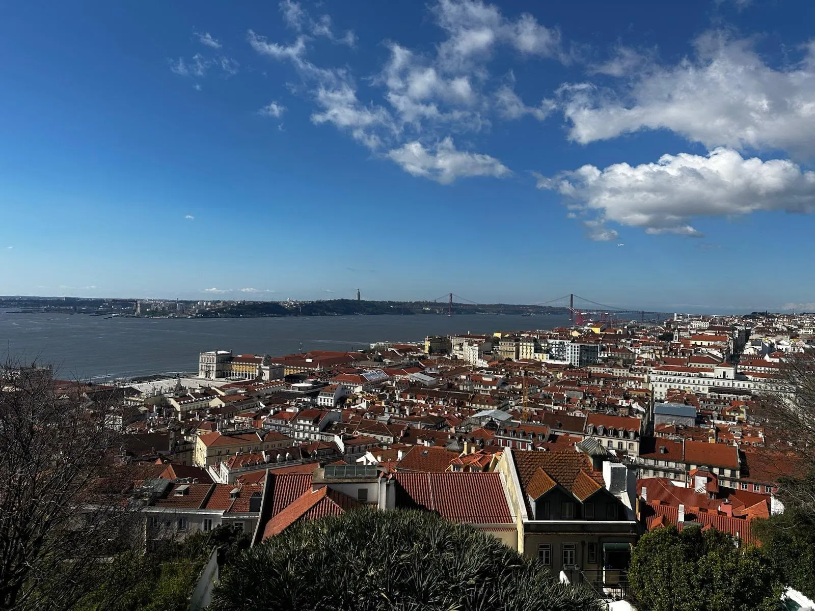 An elevated view of Lisbon's rooftops, the Tagus River, and the 25 de Abril Bridge on a sunny day.