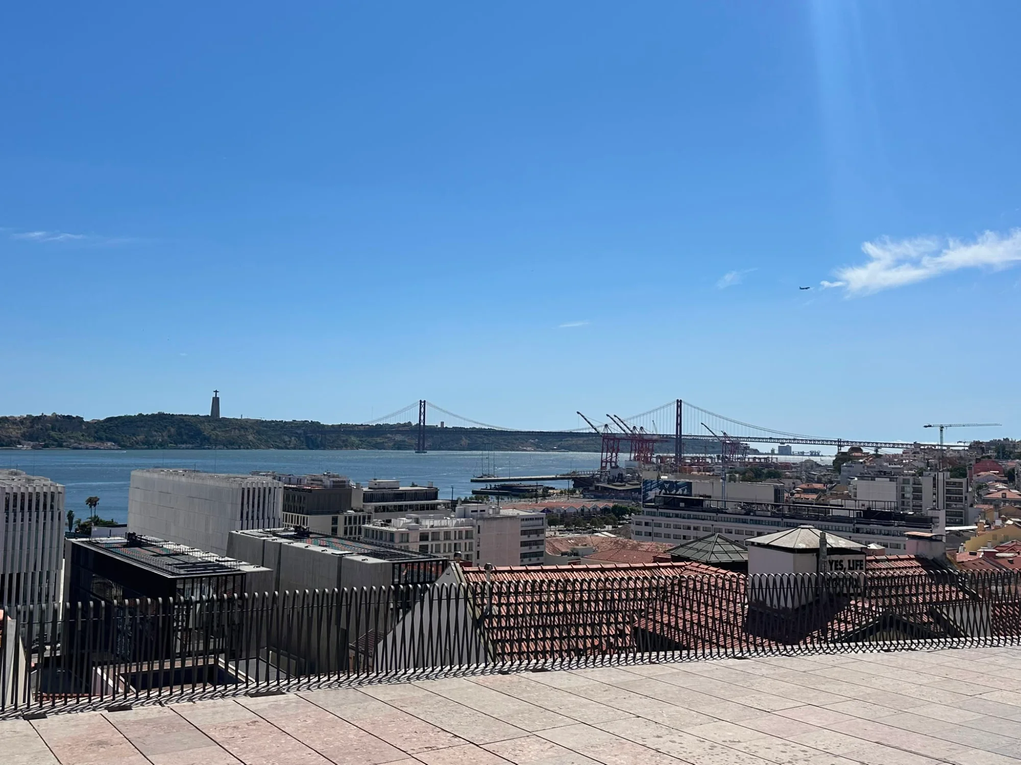 View over the rooftops of Lisbon towards the Tagus River, 25 de Abril Bridge, and Christ the King statue on a sunny day.