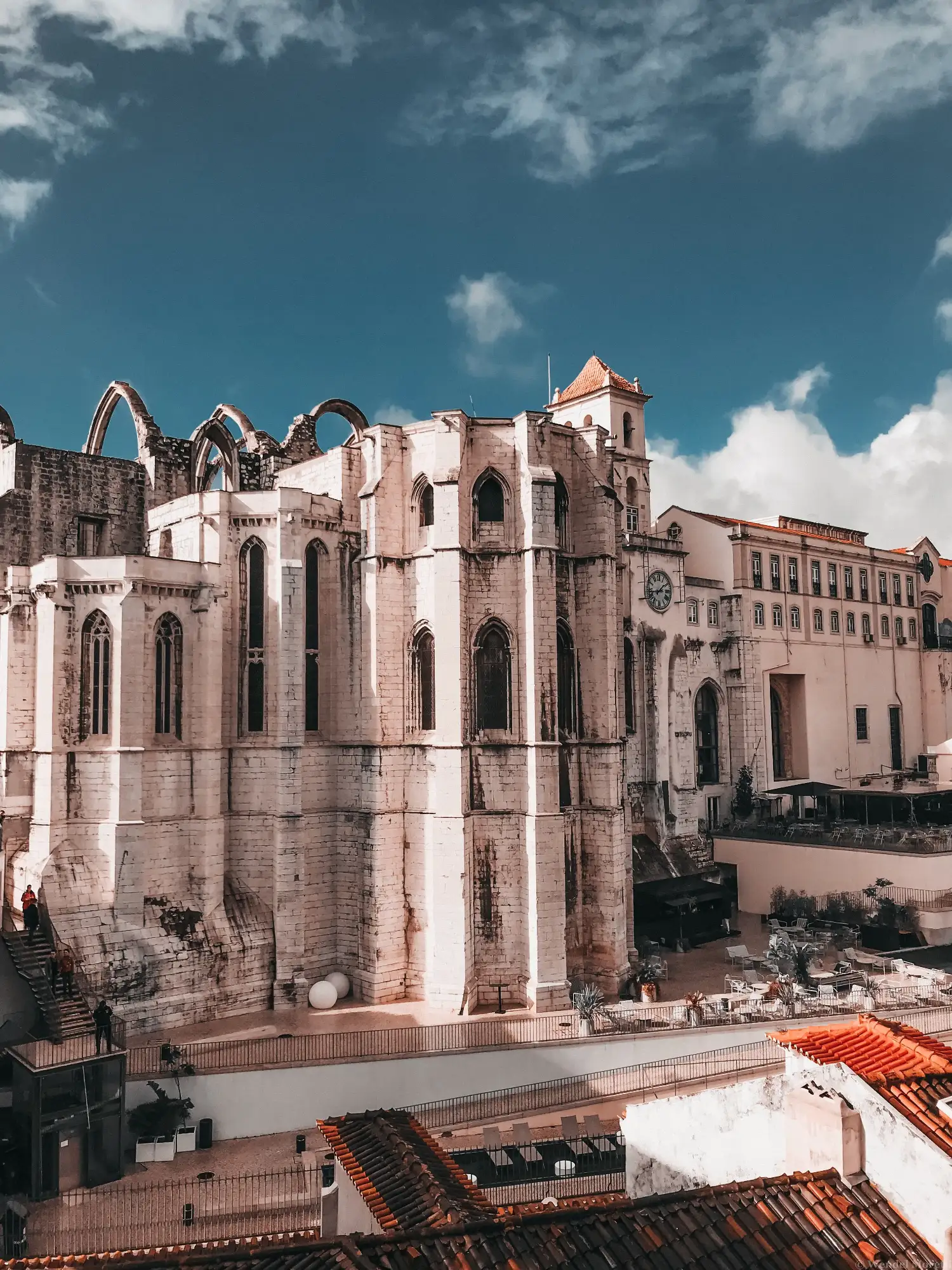 The roofless Gothic arches of the Carmo Convent in Lisbon against a blue sky with scattered clouds.