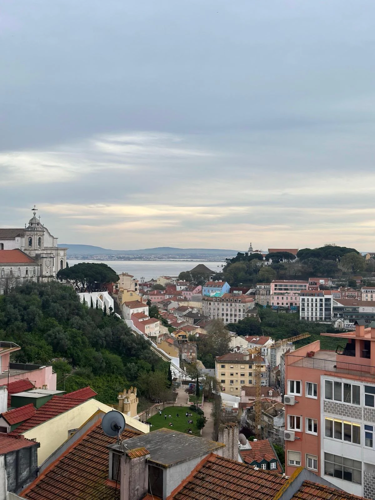 View over the rooftops of Lisbon, with a church on the left and the Tagus River in the background under a cloudy sky.