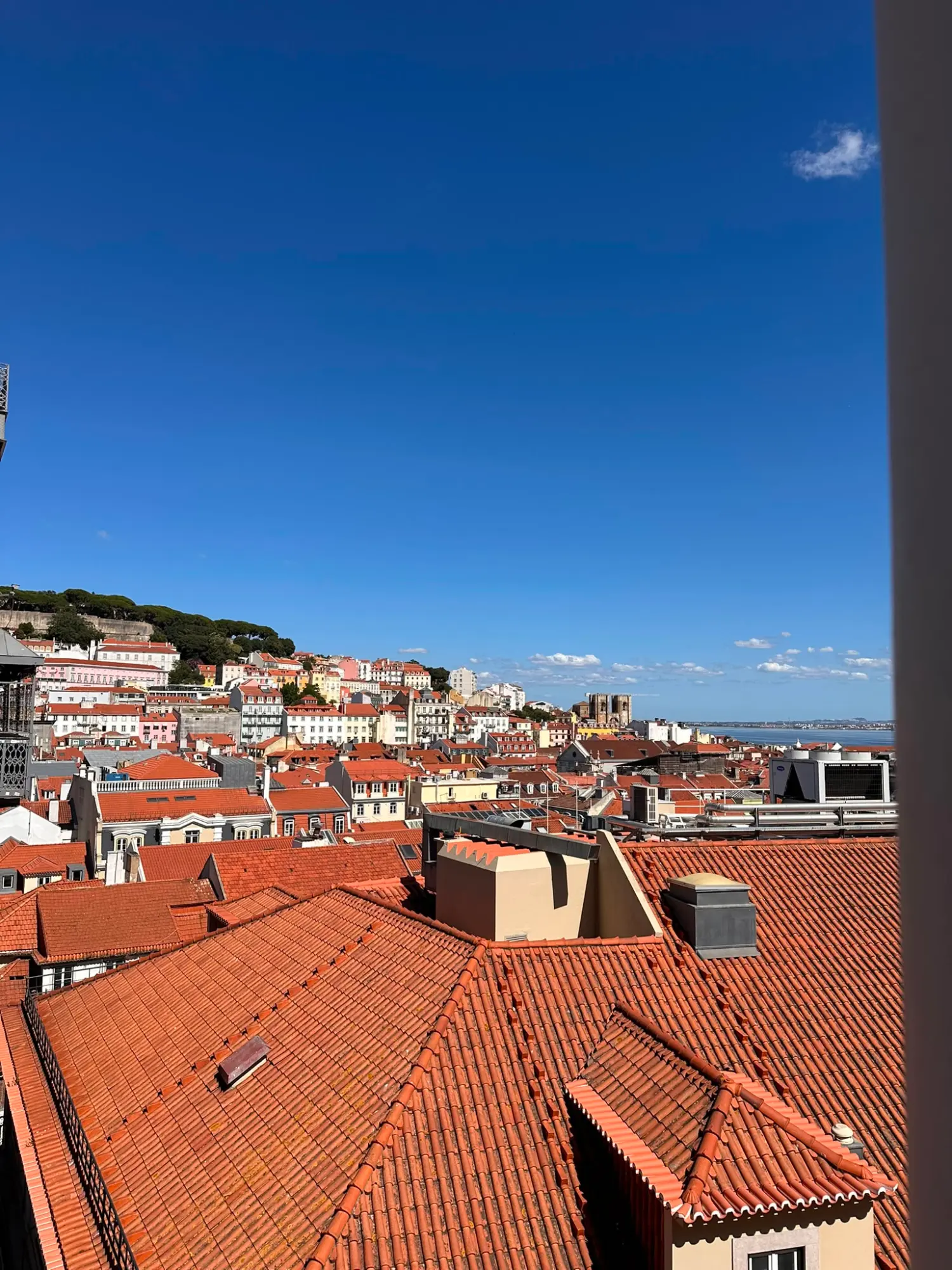 View over the red-tiled roofs of Lisbon's old town on a sunny day, with São Jorge Castle on a hill in the background.