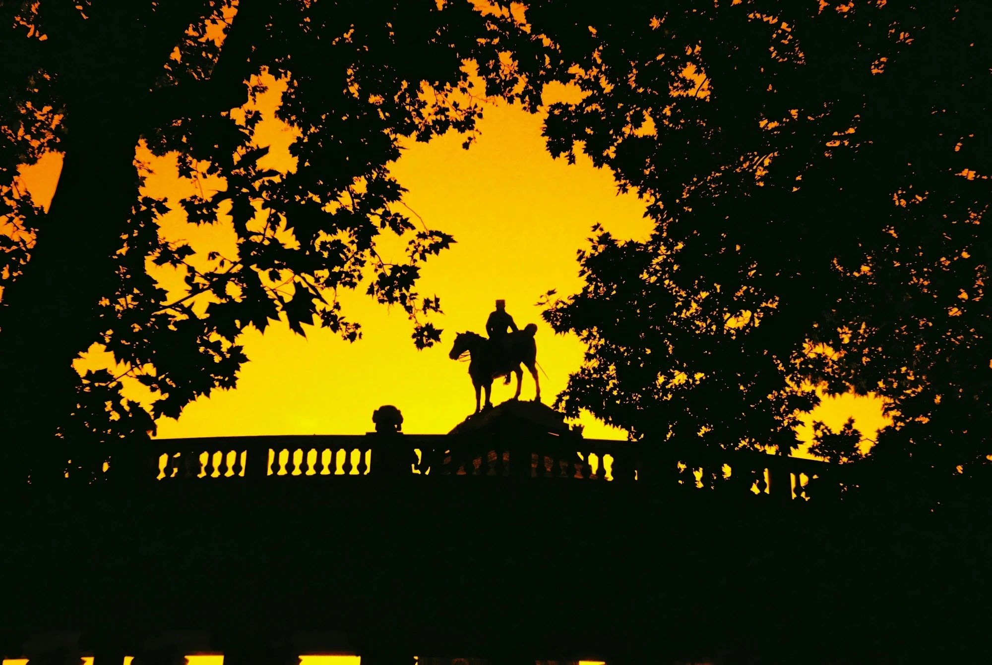 Silhouette of the equestrian statue on Wellington Arch in London against a bright orange sunset, framed by trees.