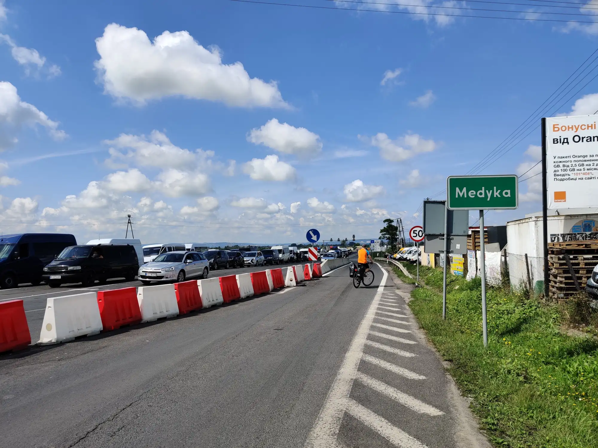 A road leading to the Medyka border crossing with a line of cars, a cyclist, and a green town sign under a blue sky.
