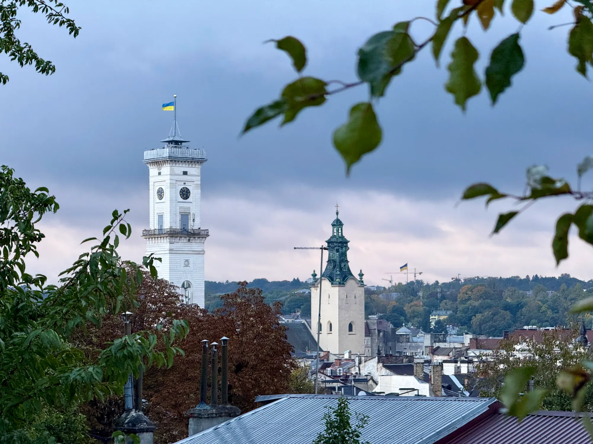 The white clock tower of Lviv Town Hall and the spire of the Latin Cathedral seen over city rooftops under a cloudy sky.