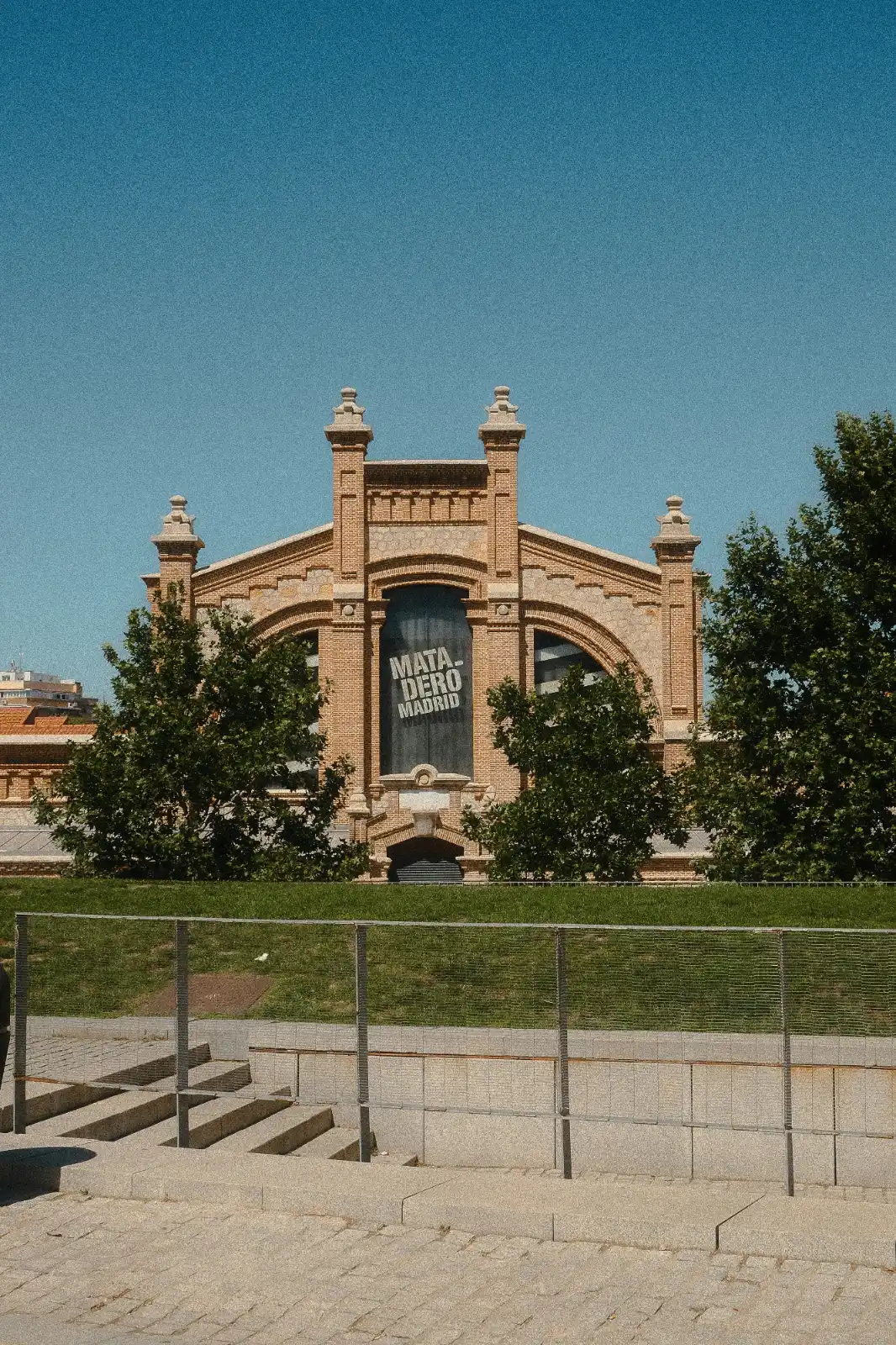 The brick exterior of the Matadero Madrid building under a clear blue sky, framed by trees in the foreground.