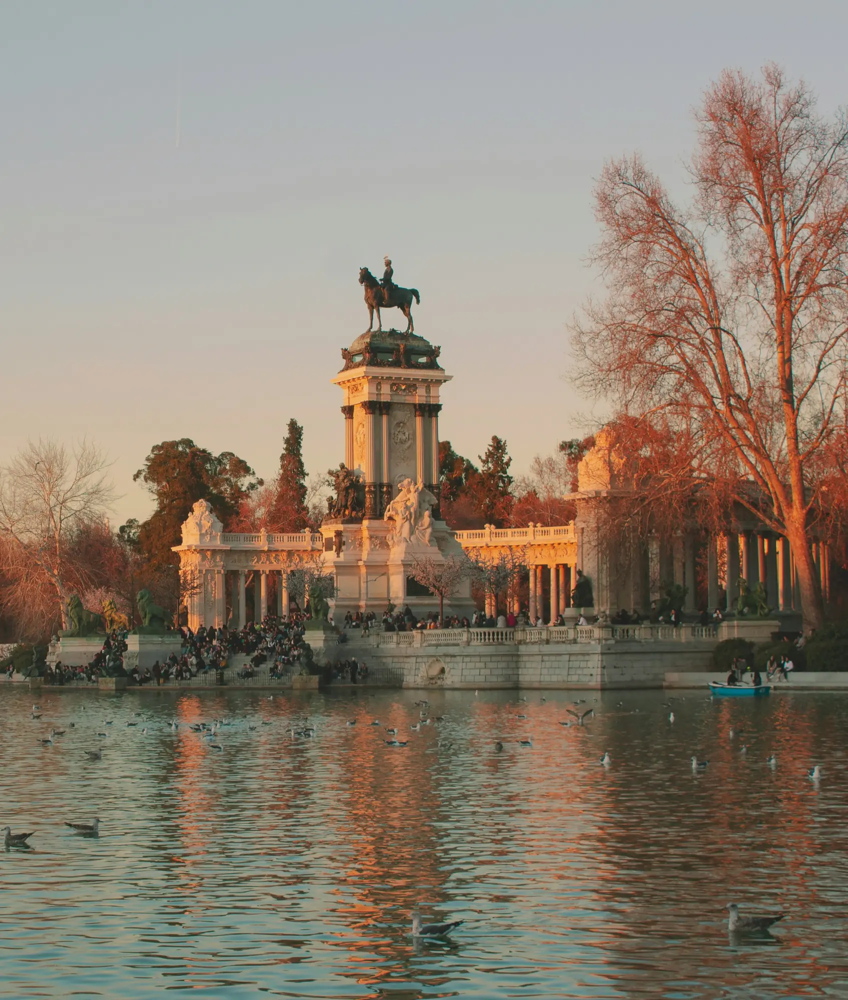 The Monument to Alfonso XII in Madrid's Retiro Park, seen from across the lake at sunset, with people relaxing on the steps.