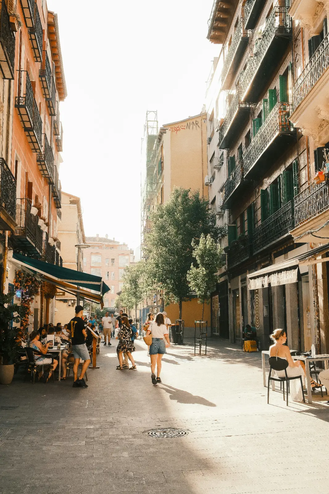 A sunny, narrow street in Madrid with pedestrians and people sitting at outdoor cafes.