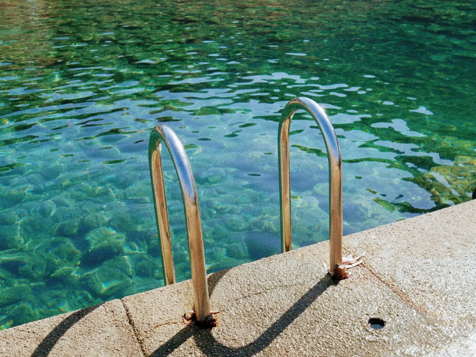 A metal ladder with curved rails leads from a concrete pier into clear turquoise water with a rocky bottom.