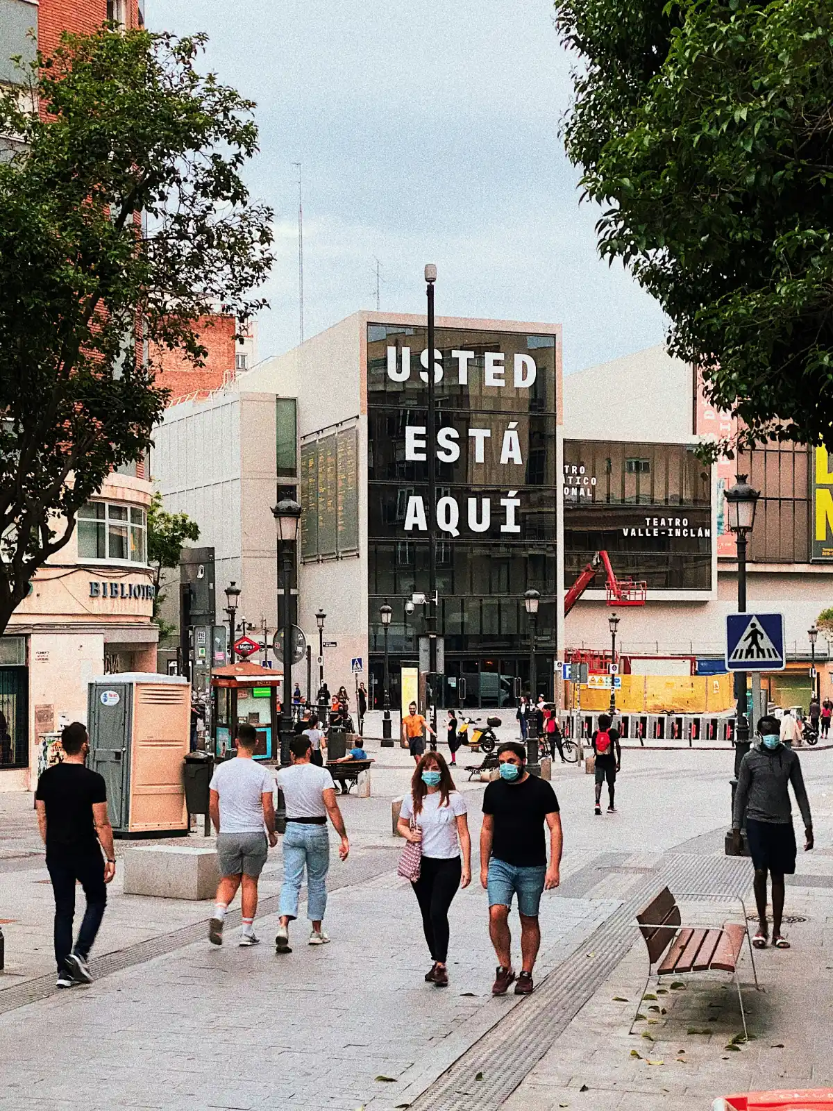Pedestrians on a city street in front of the Teatro Valle-Inclán, which has 'USTED ESTÁ AQUÍ' on its facade.