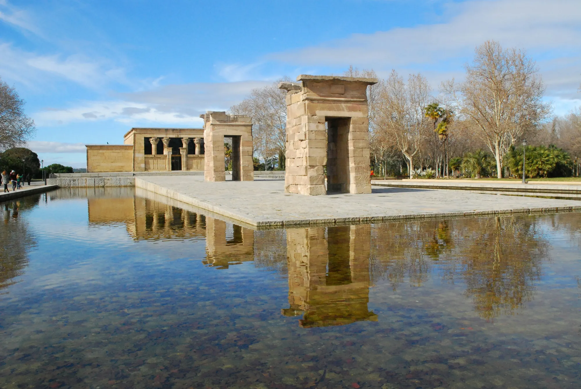 The ancient Egyptian Temple of Debod and its gateways reflected in a clear pool of water under a blue sky.