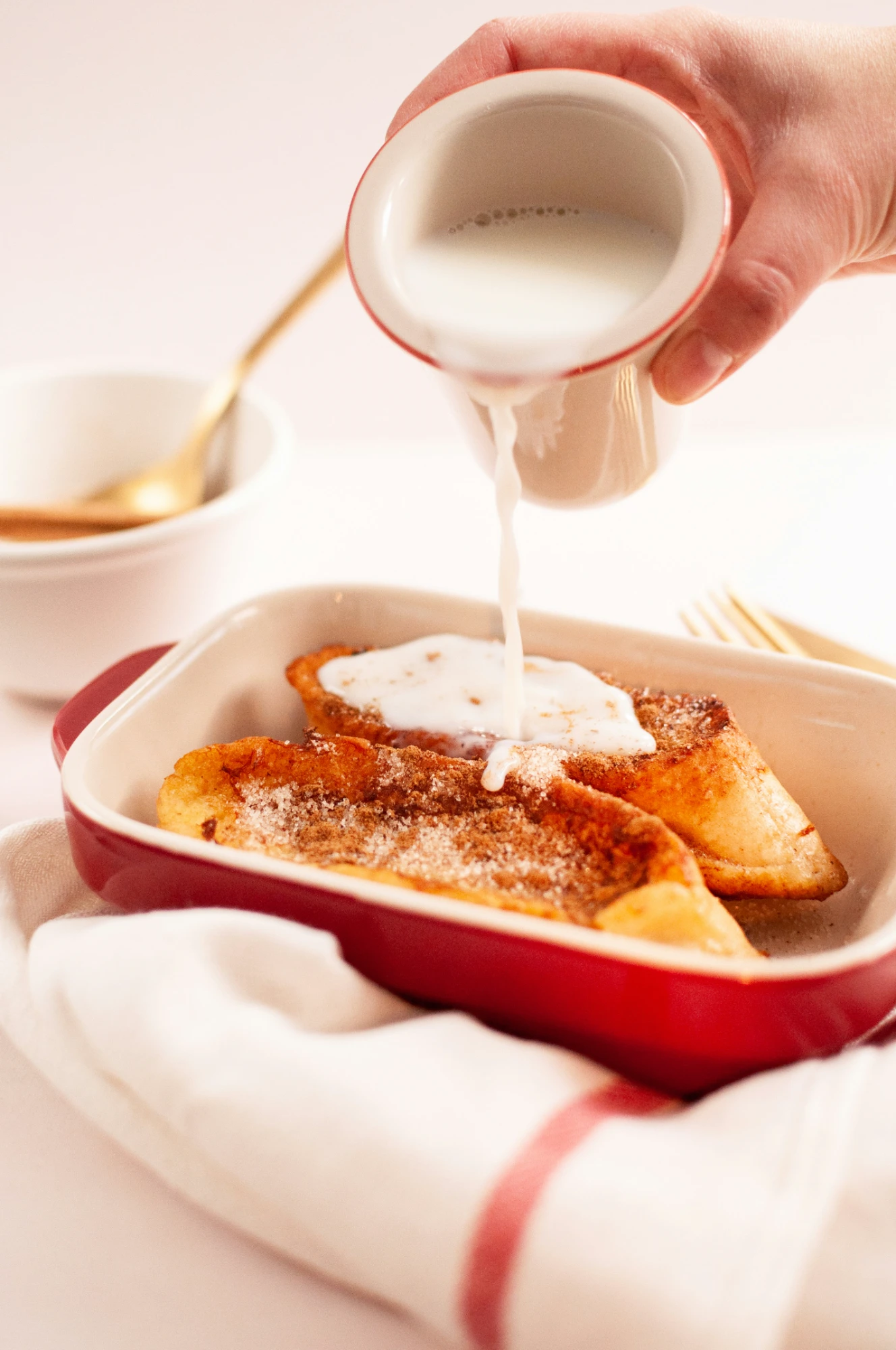 A hand pours milk over two slices of torrijas, a traditional Spanish dessert, in a small red ceramic dish.