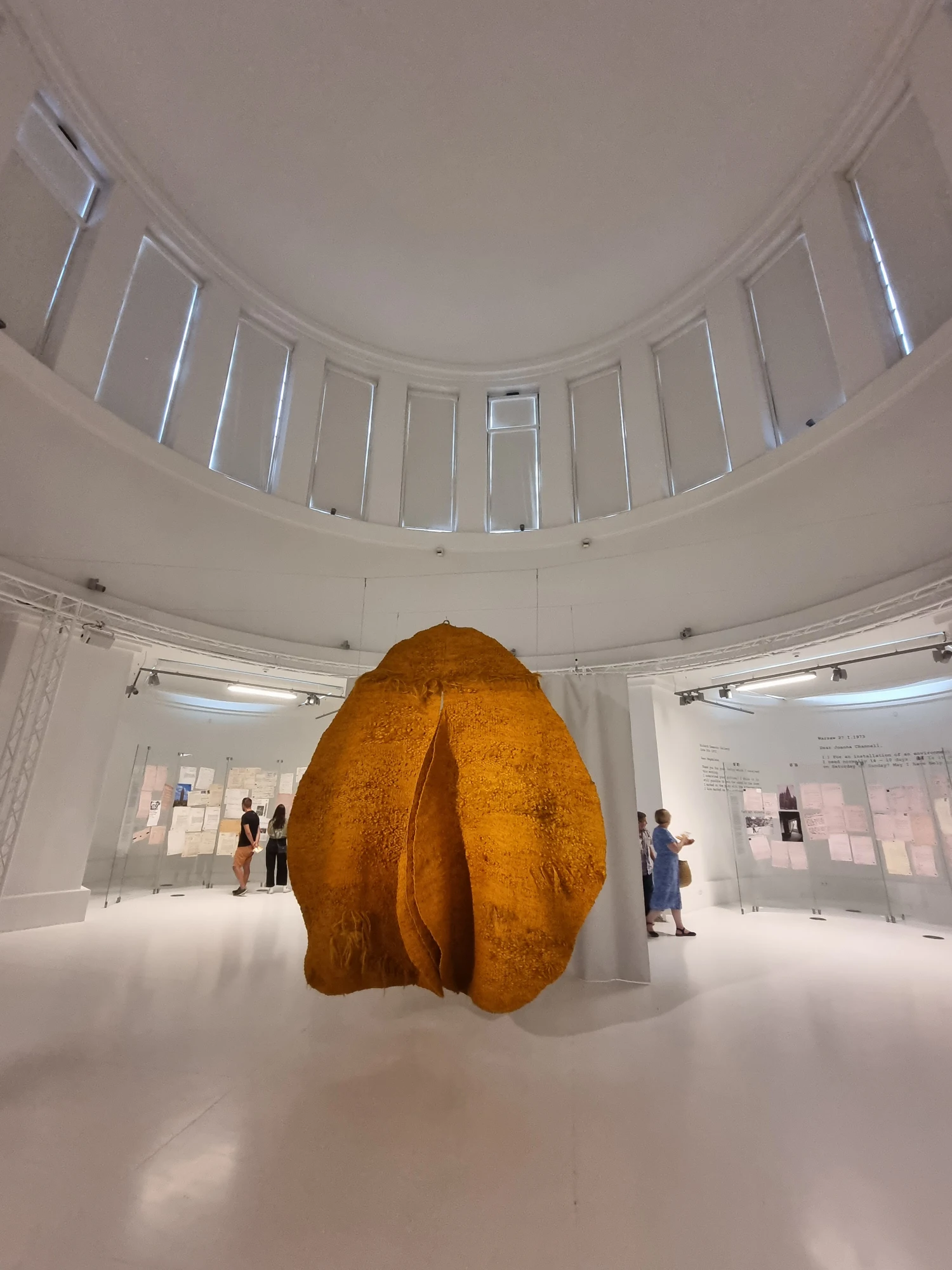 A large, yellow, abstract sculpture hangs in the middle of a circular, white museum gallery with a high domed ceiling.
