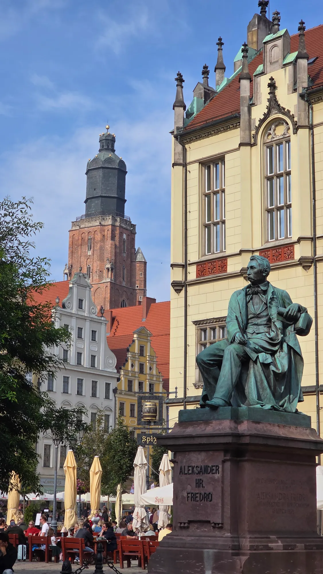 A bronze statue of Aleksander Fredro in Wrocław's Market Square, with historic buildings and a church tower in the backgro...