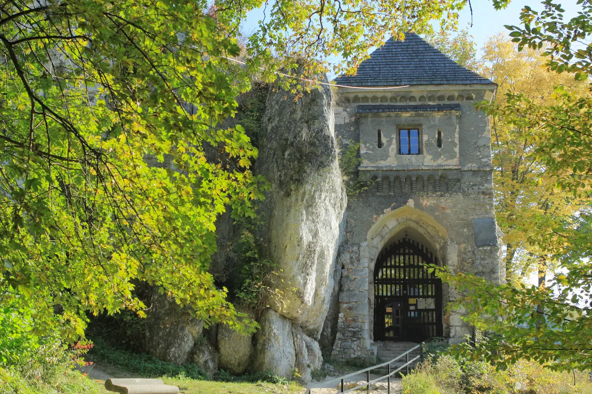 A medieval stone gatehouse with a dark wooden gate, built into a limestone cliff and surrounded by early autumn trees.