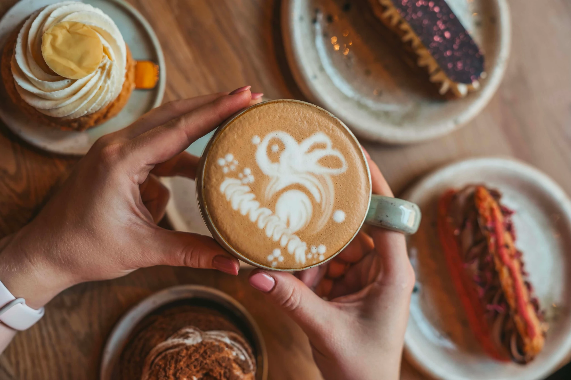 Dłonie trzymające filiżankę kawy z latte art, w otoczeniu ciast i wypieków na drewnianym stole w kawiarni Chmiel Kawę.