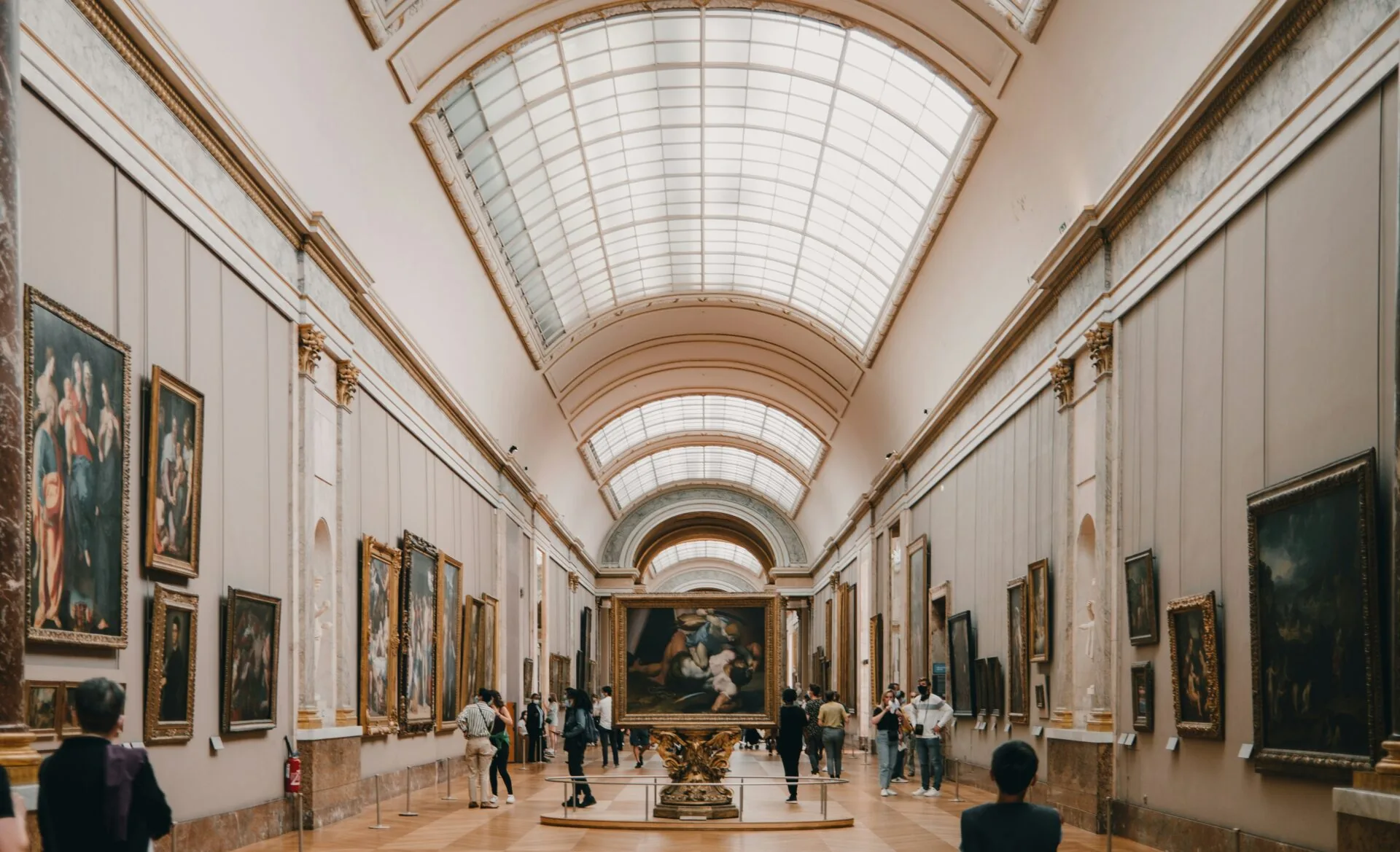 Wielka galeria sztuki w Paryżu z licznymi obrazami na ścianach i sklepionym szklanym sufitem. Zwiedzający oglądają dzieła.