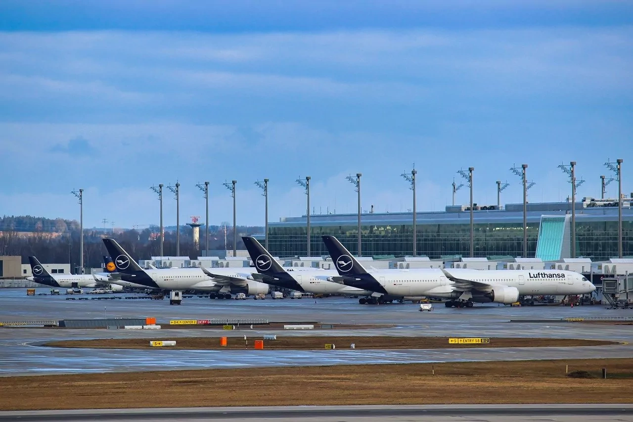Several Lufthansa passenger airplanes parked at gates at a large airport terminal under a partly cloudy sky.