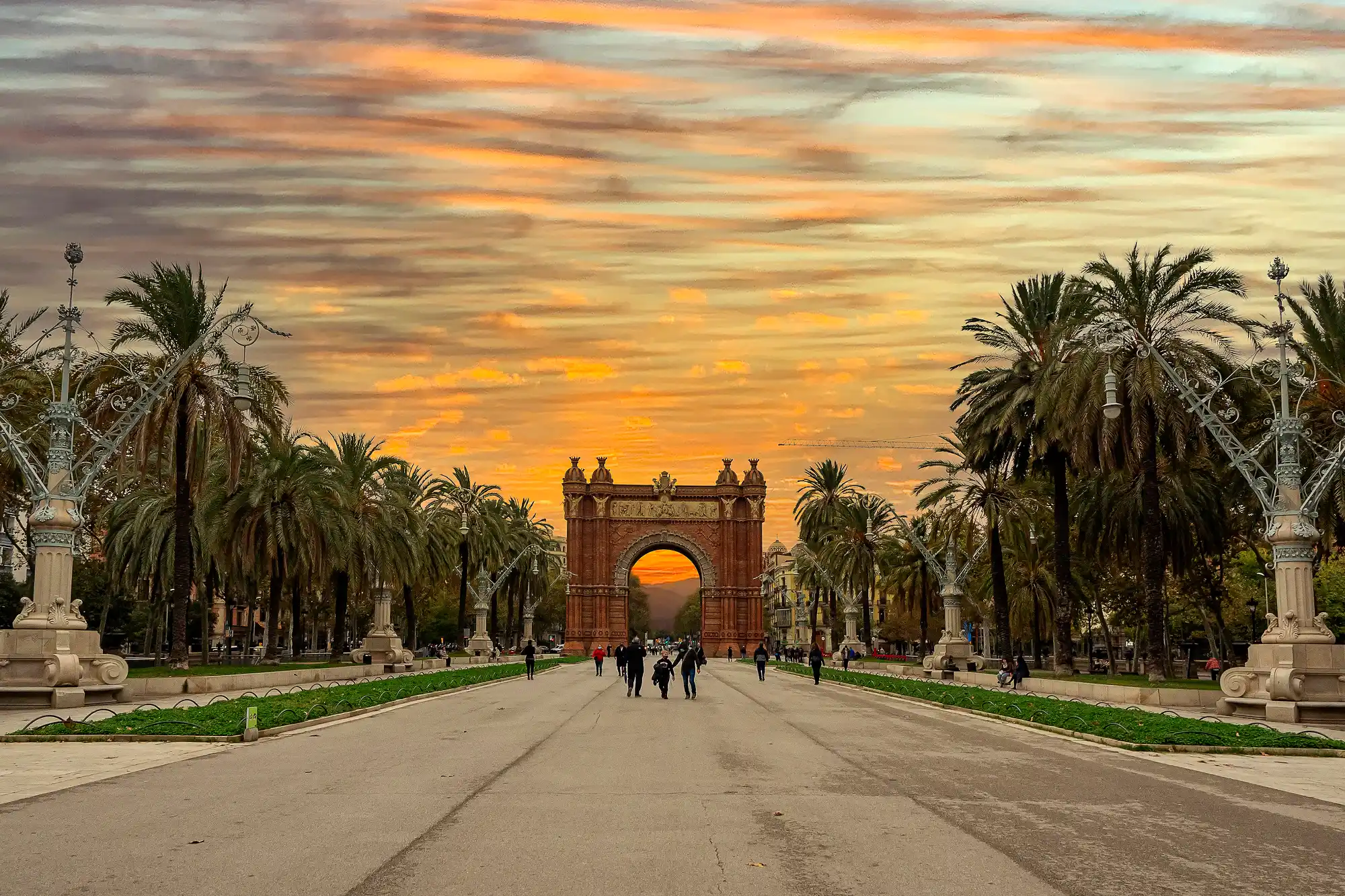 The Arc de Triomf in Barcelona at the end of a palm-lined promenade, under a vibrant orange sunset sky.