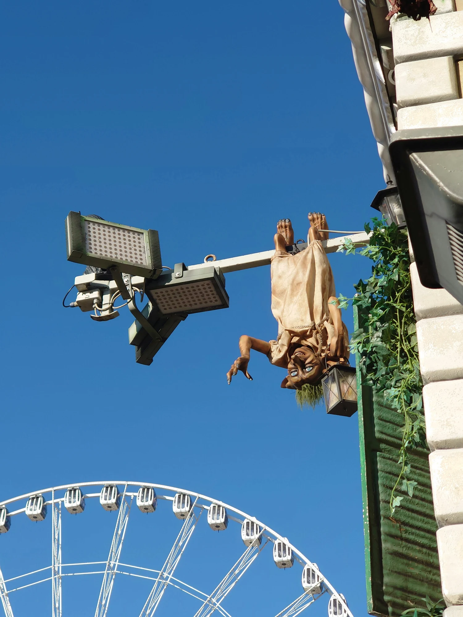 Eine koboldähnliche Figur hängt kopfüber an einer Stange und hält eine Laterne, mit einem Riesenrad im Hintergrund.