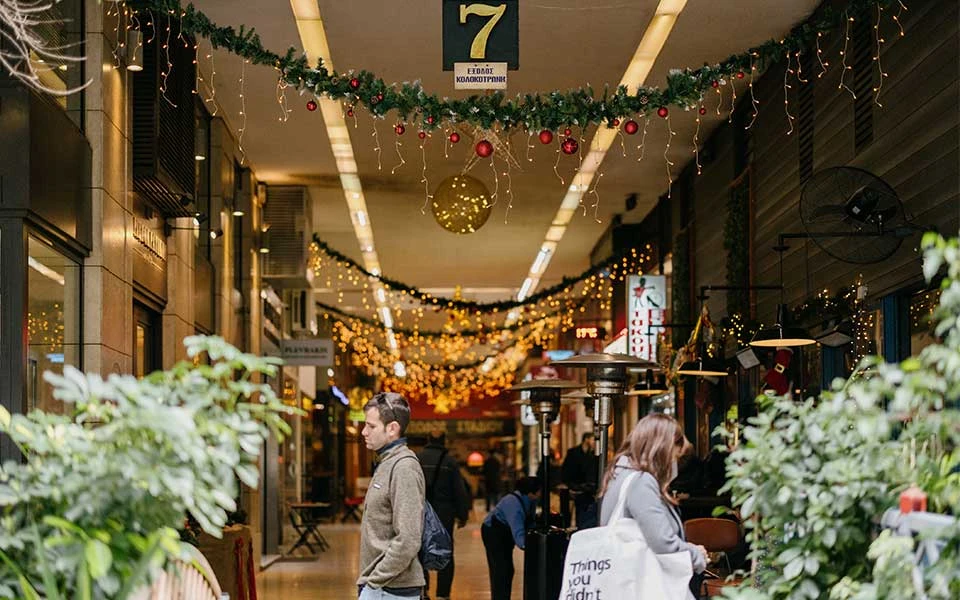 Passanten in einer Athener Einkaufspassage mit festlicher Weihnachtsbeleuchtung und Tannengirlanden.