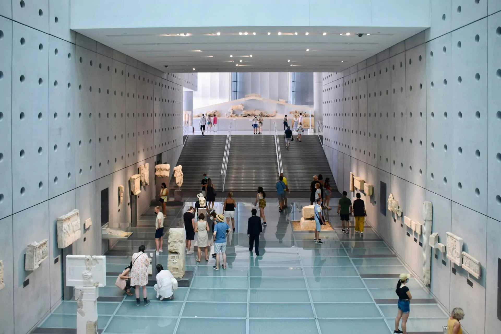 Visitors walking up the wide stairs and glass ramp inside the modern Acropolis Museum in Athens.
