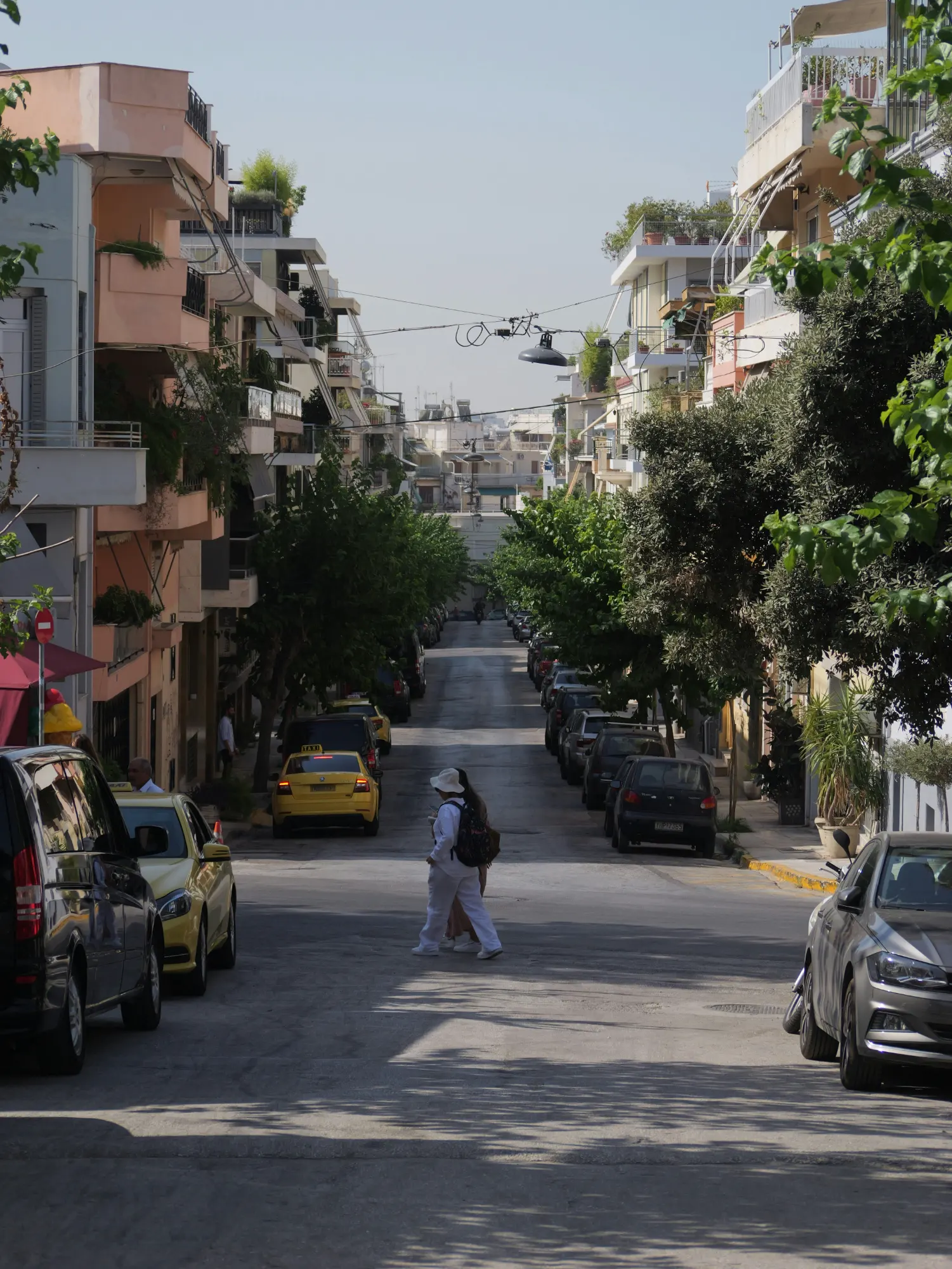 View down a residential street in Koukaki, Athens, lined with apartment buildings, parked cars, and trees.