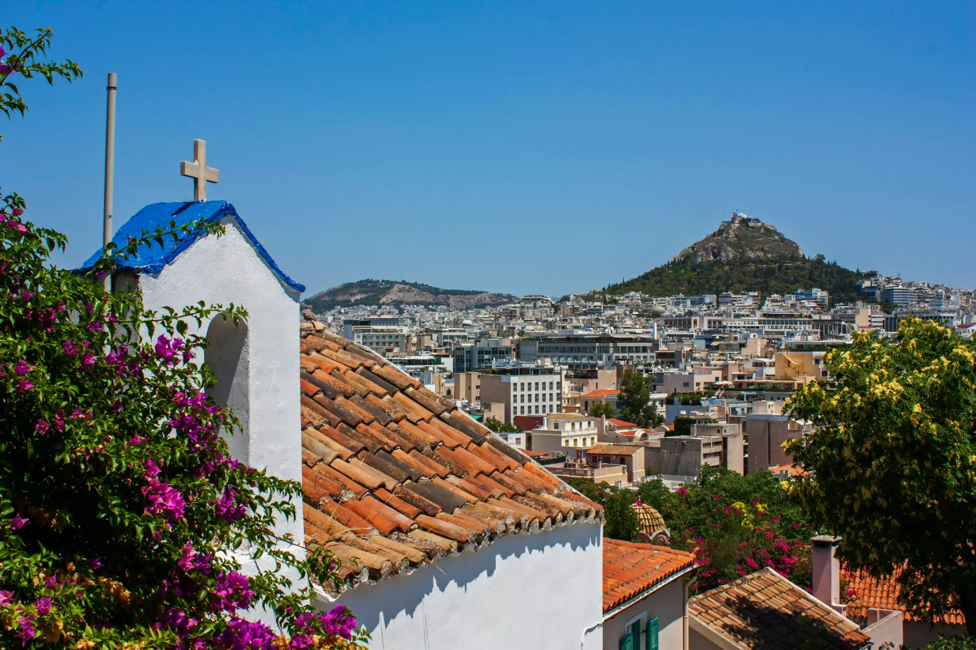 Weiße Kapelle mit blauem Dach und Kreuz, Bougainvillea, rote Ziegeldächer, Athener Stadtansicht und Lykabettus-Hügel unter bl