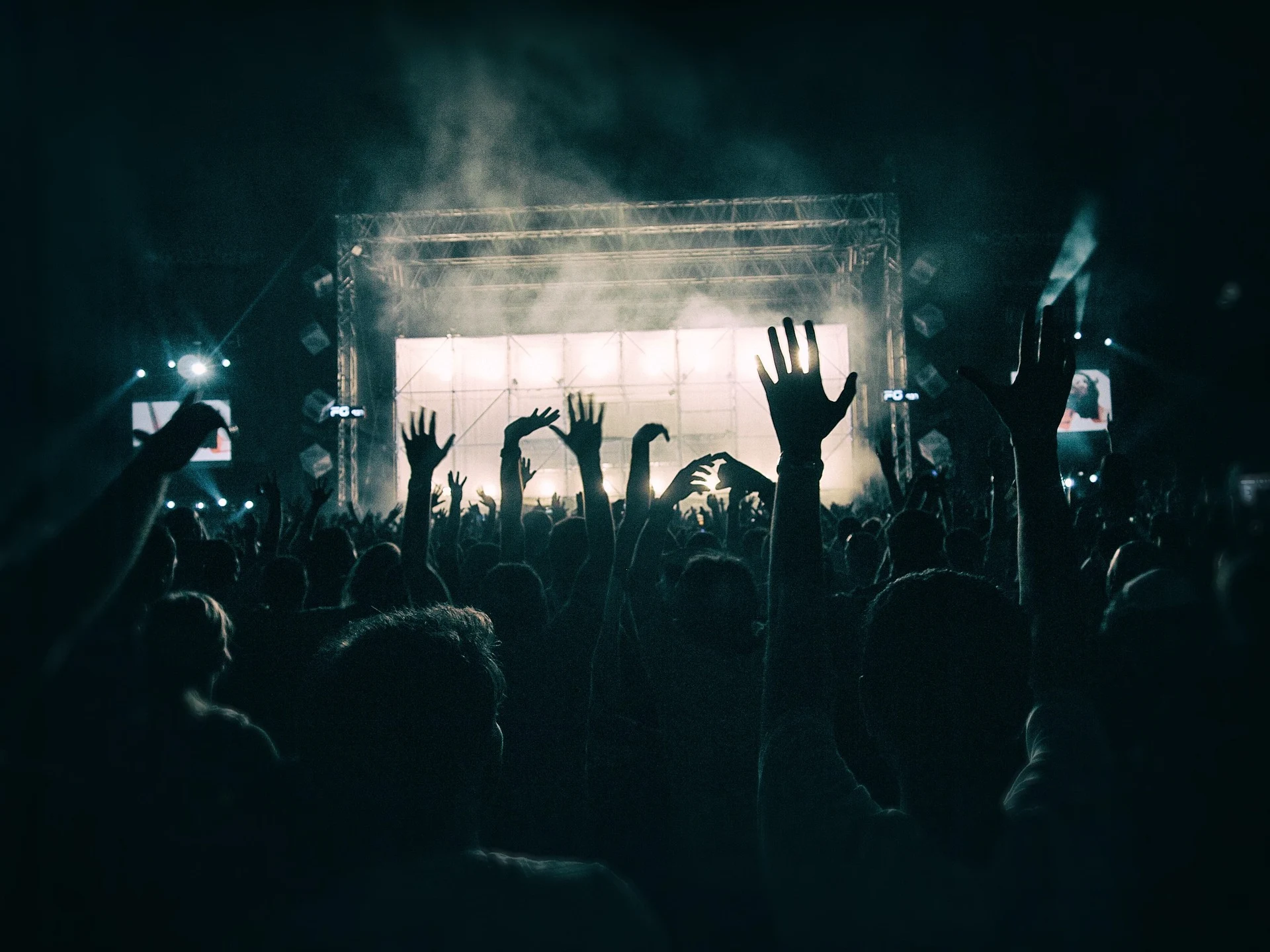 Silhouetted audience with raised hands at a nighttime concert, light and smoke from a stage visible in the background.