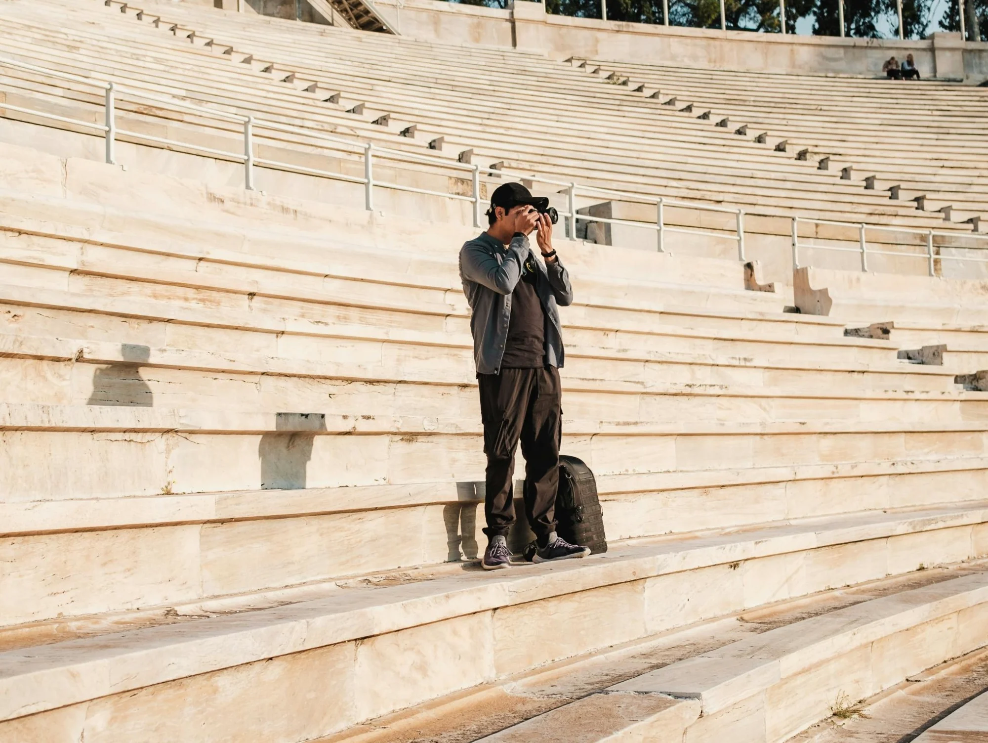 Man in a cap and grey jacket taking a picture from the light marble steps of a large stadium, with empty rows behind.