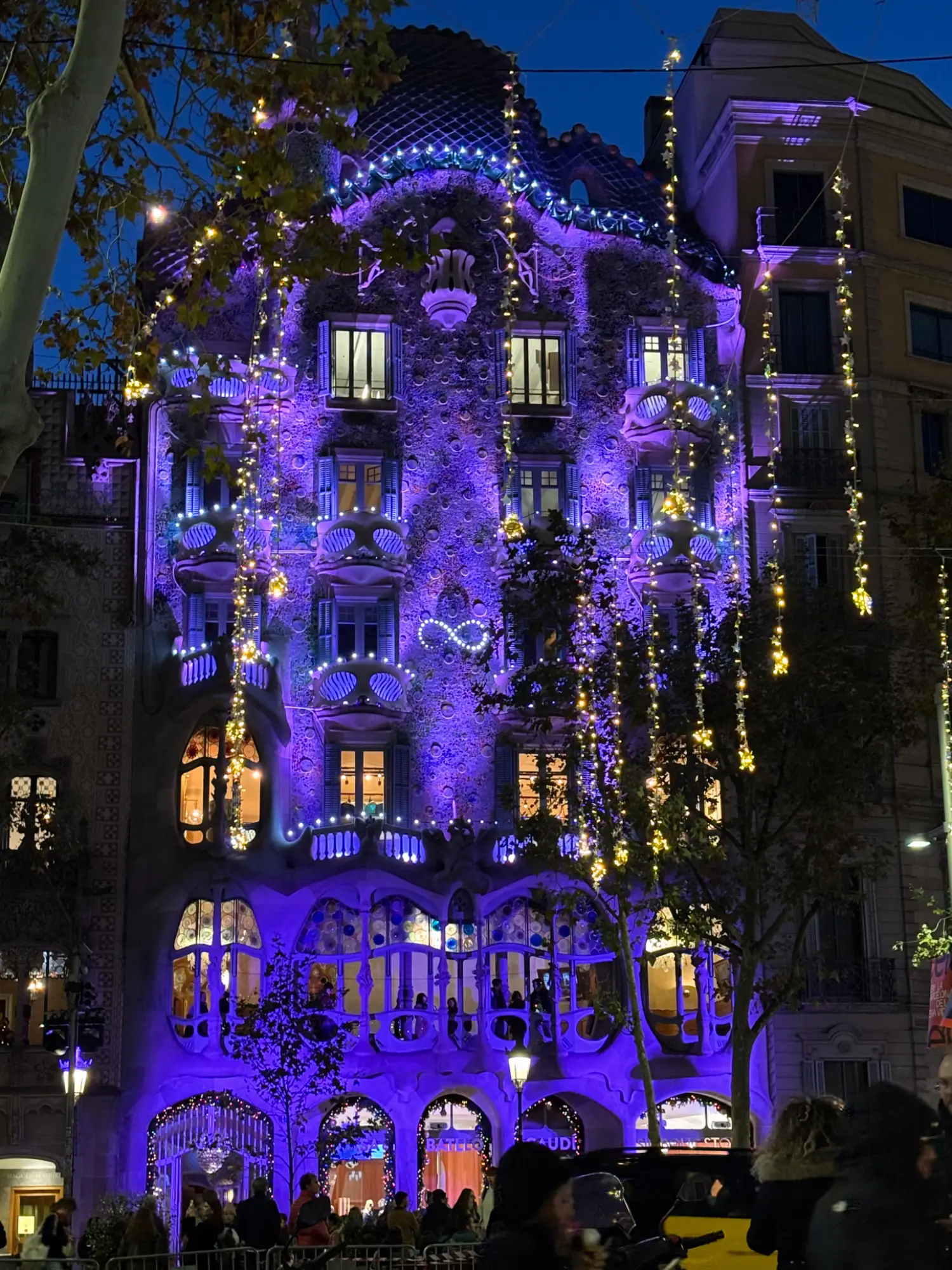 Casa Batlló illuminated in purple at night, adorned with cascading yellow string lights and trees, with people on the street.