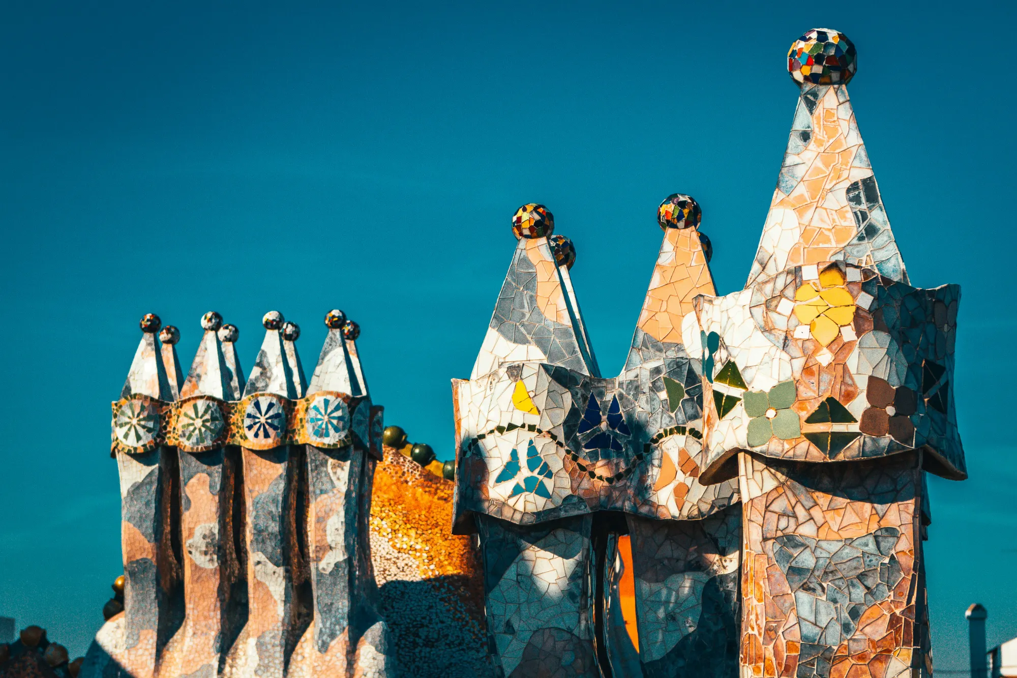 Close-up of ornate, colorful mosaic-covered chimneys and spires with spheres on top against a clear blue sky.