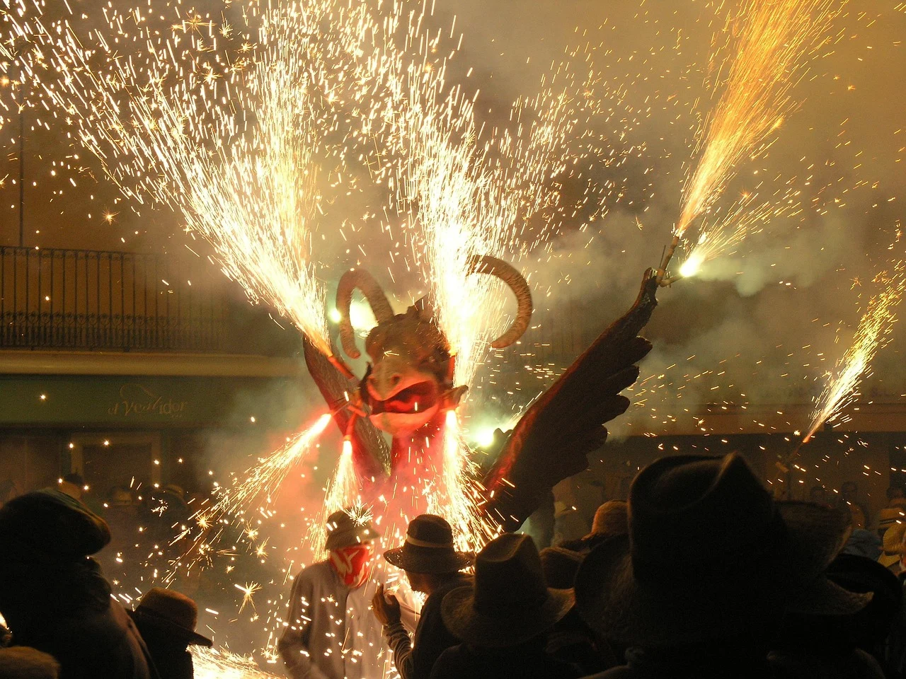 Gran figura de dragón alado con cuernos que emite chispas y fuegos artificiales en un festival nocturno, observado por person