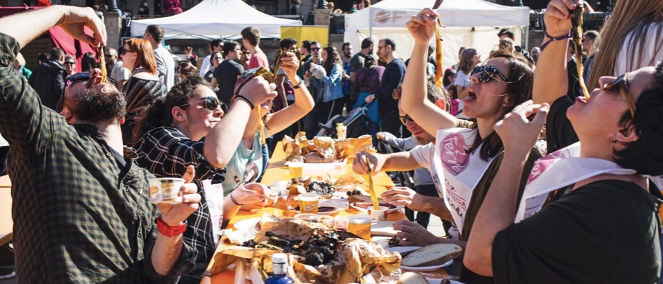 People at a calçotada festival in Barcelona eating grilled calçots while wearing bibs. Many charred onion skins on the table.