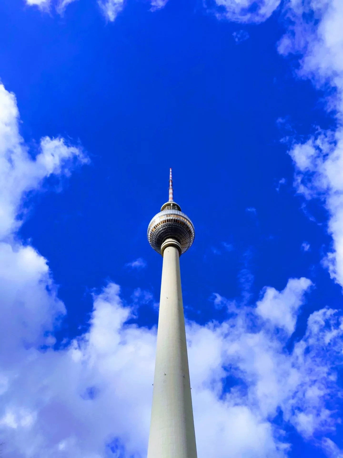 The Berlin TV Tower, Fernsehturm, viewed from below against a bright blue sky with scattered white clouds.