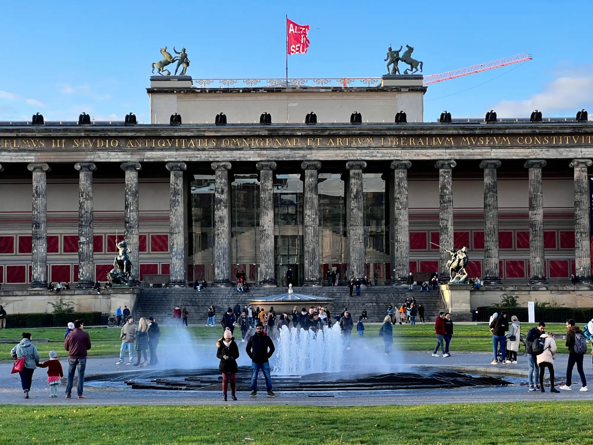 The classical facade of the Altes Museum with columns, roof statues, a red flag, and a fountain in Lustgarten, with people wa