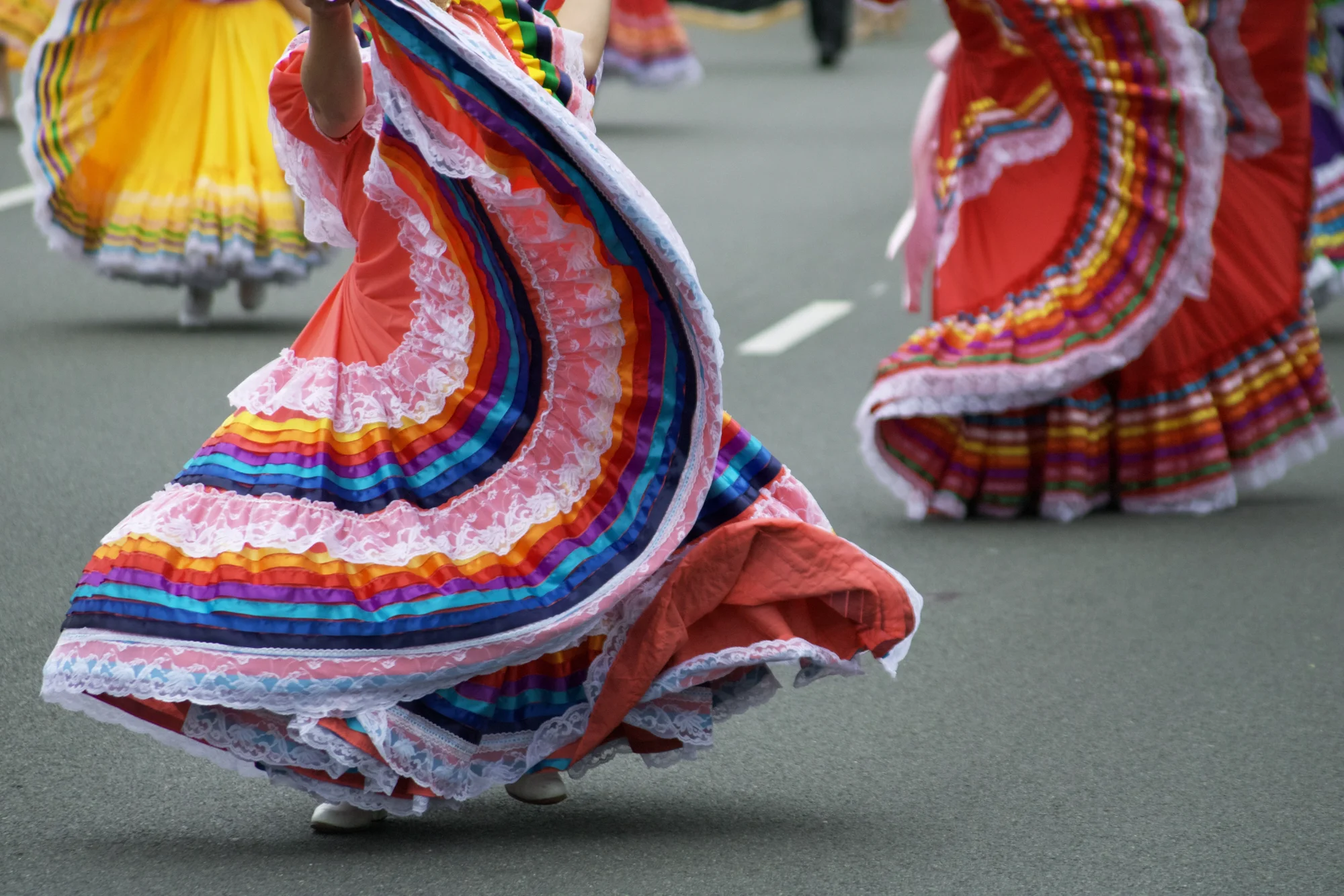 Dancers in colorful, layered dresses with rainbow ribbons and white lace during a street parade.