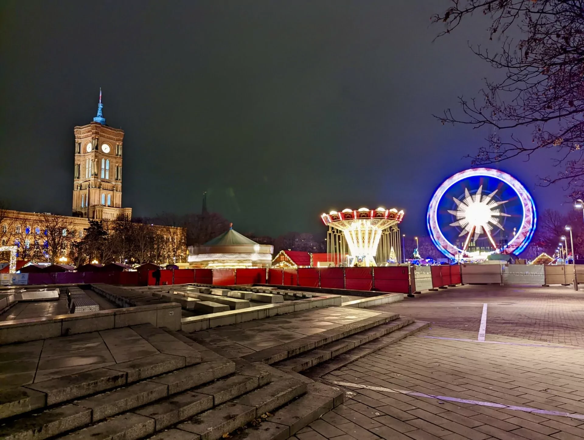 Night view of a Ferris wheel, carousel, and market stalls with the illuminated Rotes Rathaus clock tower in Berlin.