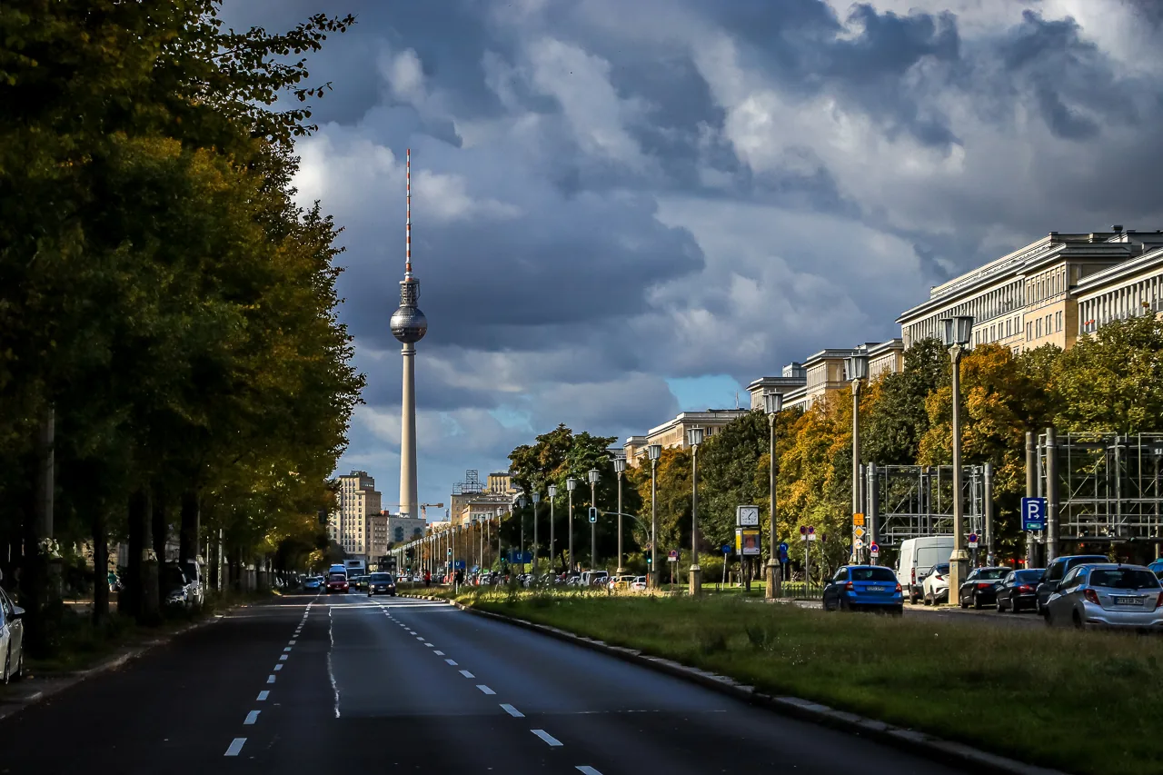 Street view of the Berlin TV Tower (Fernsehturm) on a cloudy day, with cars on the road and autumn trees.