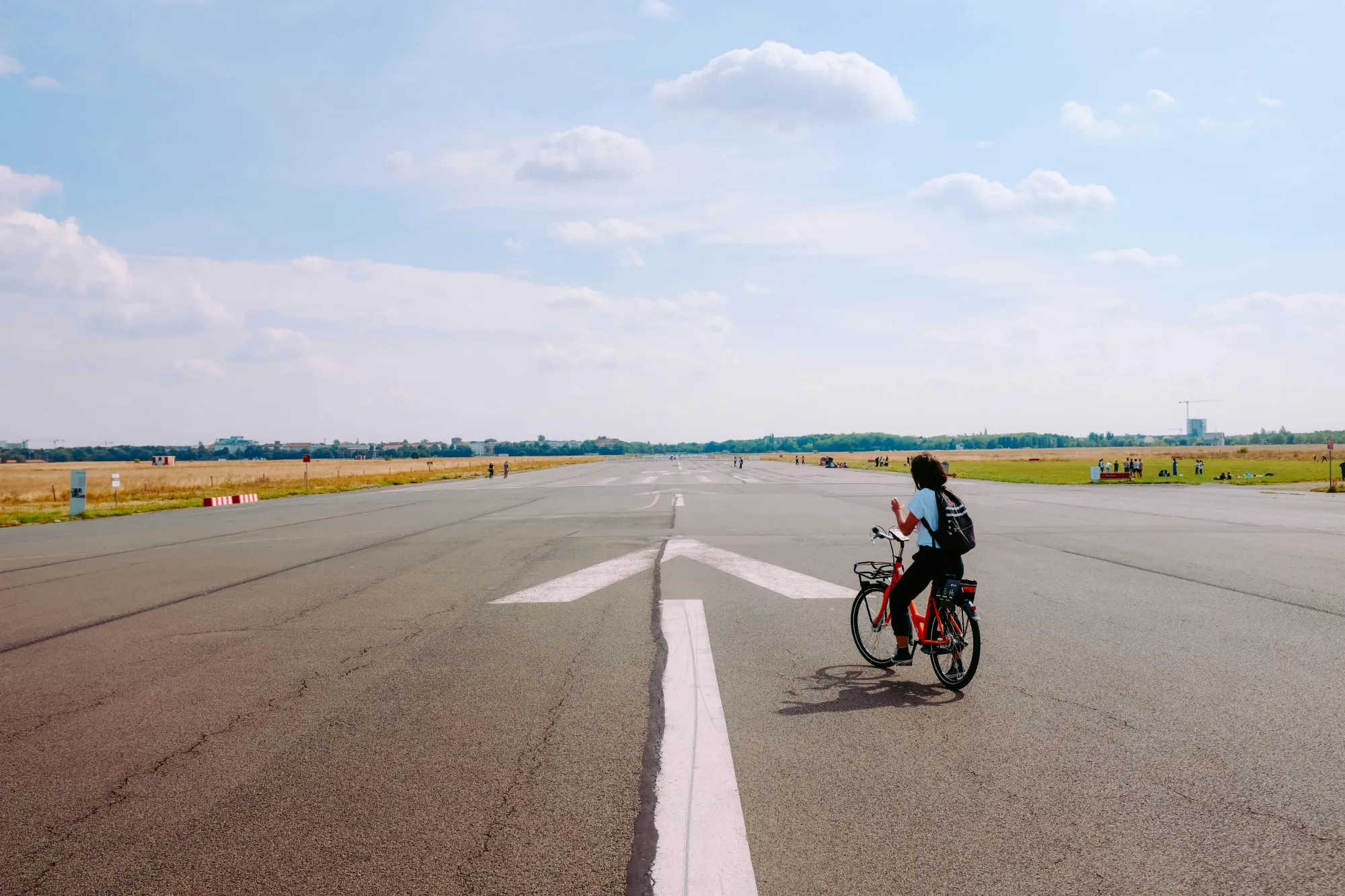 Persona en bicicleta roja en la pista asfaltada de Tempelhofer Feld en Berlín, con campos y edificios a lo lejos.