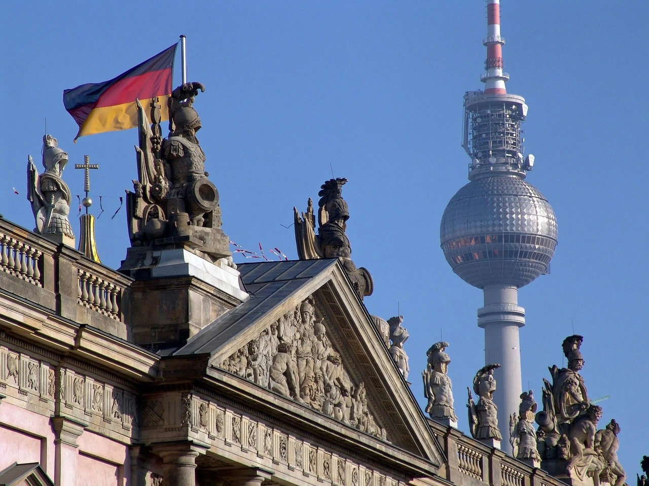 Zeughaus roof statues and German flag in the foreground with the iconic Berlin TV Tower rising behind against a blue sky.