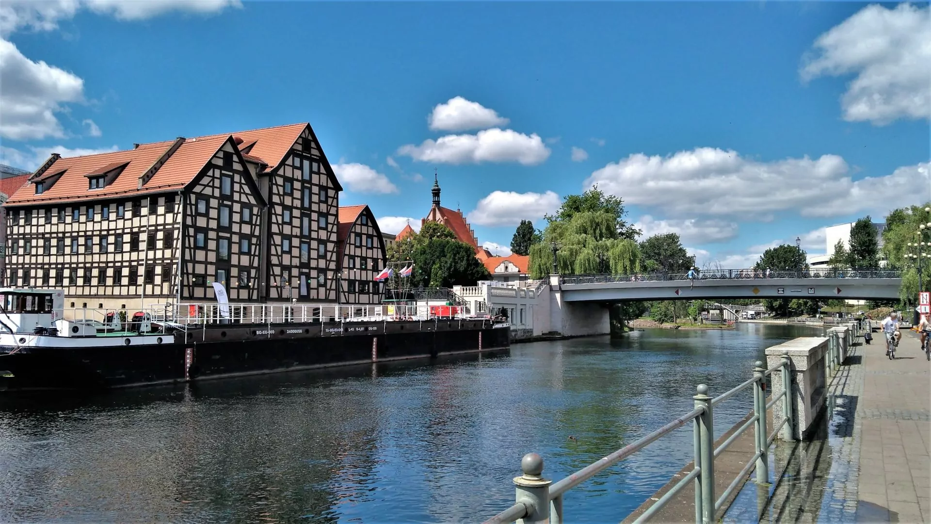 Graneros de entramado de madera junto al río Brda, con puente, barcaza y peatones en un día soleado.