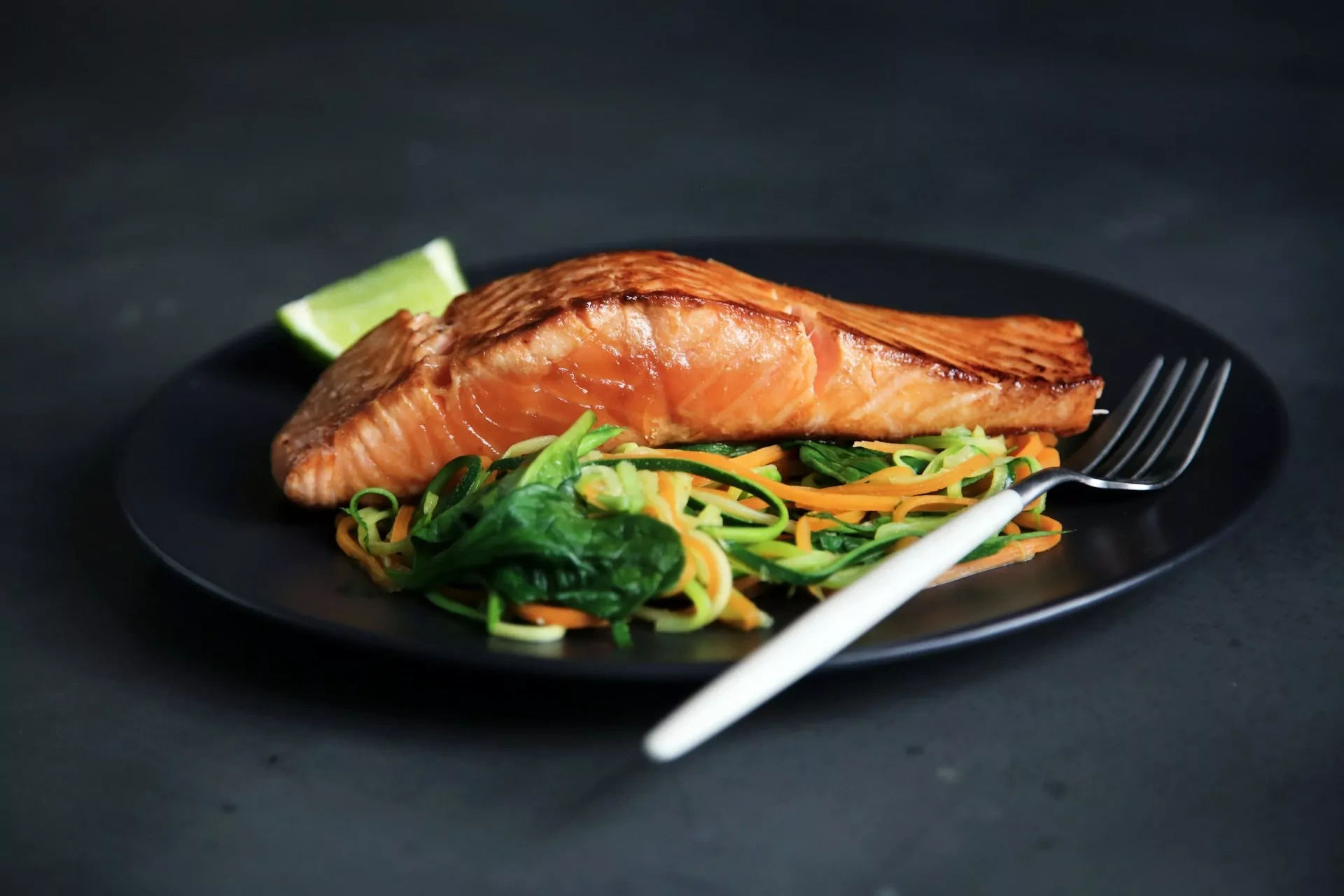 Cooked salmon fillet on a bed of spiralized carrots, zucchini, and spinach, with a lime wedge and a fork on a dark plate.