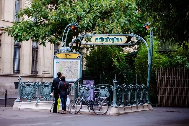 Wejście do stacji paryskiego metra Cité w stylu Art Nouveau, z dwiema osobami oglądającymi mapę i zaparkowanym rowerem.