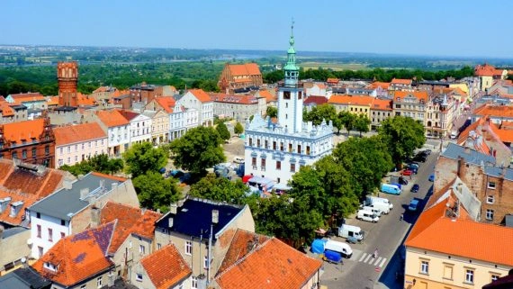 Aerial view of a historic European town square with a white town hall, green-spired tower, red roofs, and trees.