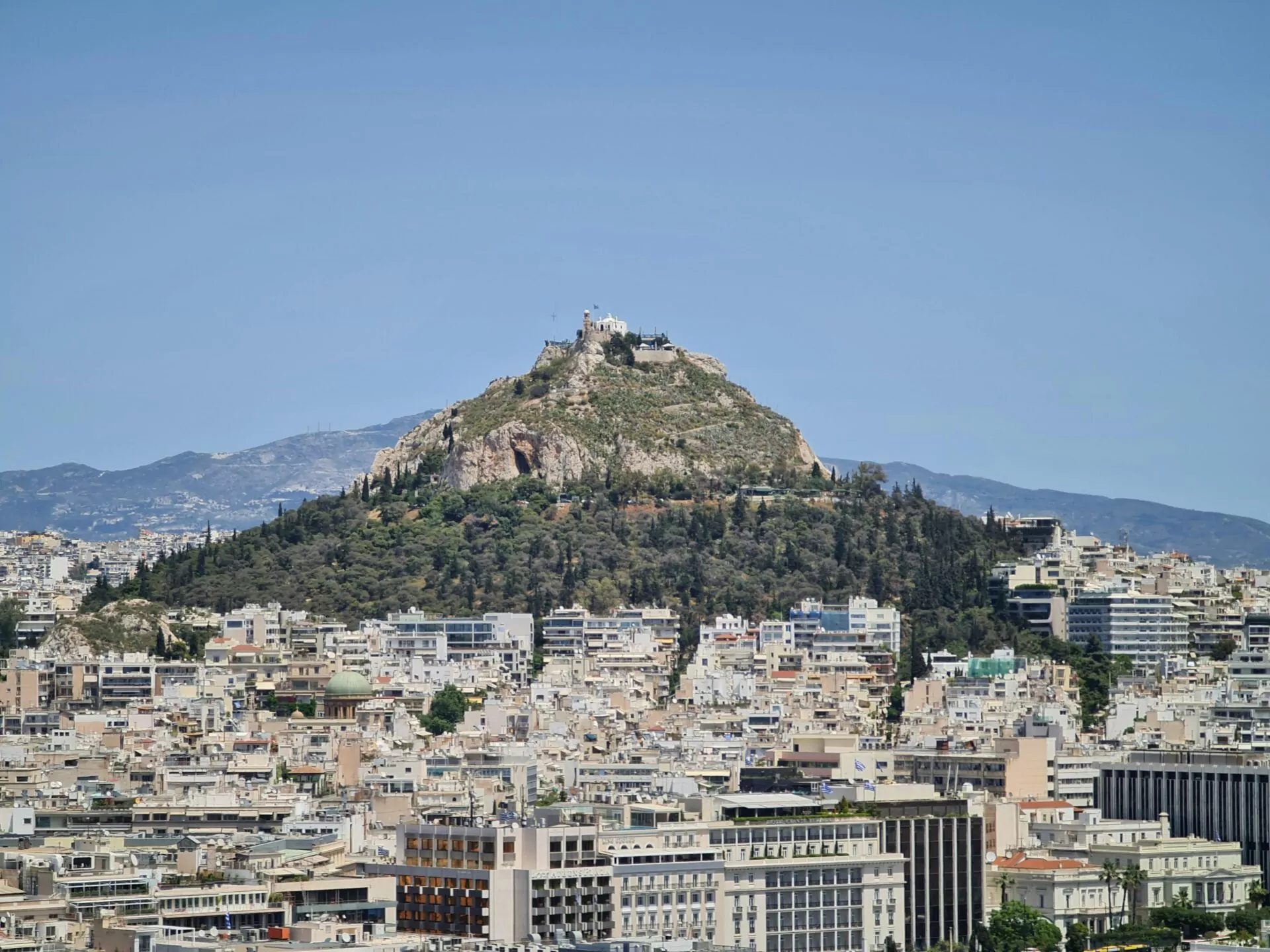 Panoramic view of Athens cityscape with Lycabettus Hill crowned by a white chapel, under a clear blue sky.