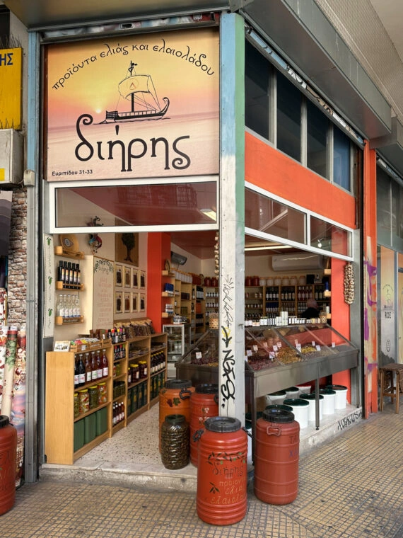 Street view of an Athens shop selling olive products and oil, featuring red barrels outside and shelves packed with jars insi
