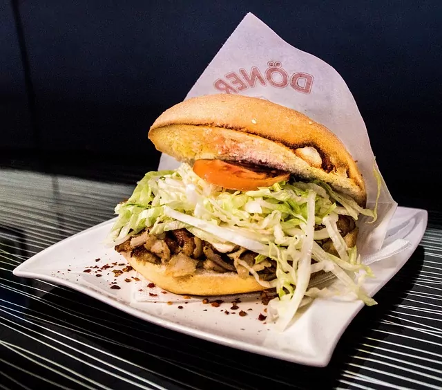 A Döner Kebab in a bun with meat, shredded cabbage, and a slice of tomato, served on a white plate. Paper wrapper with "Döner