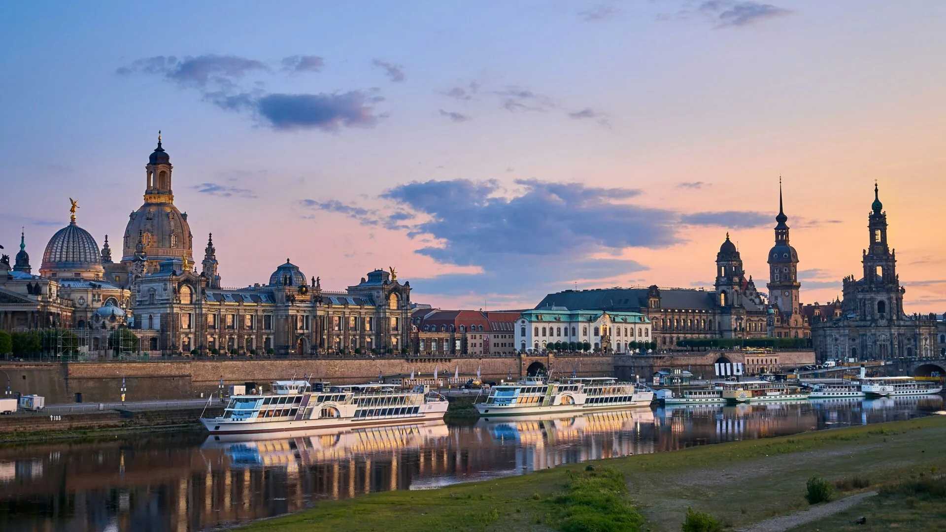 Historic Dresden skyline with Frauenkirche and Hofkirche reflected in the Elbe River at sunset, with riverboats docked.